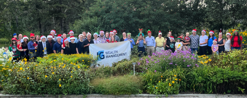 members of the IG Wealth Management team and their families stand behind a bed of colourful perennial flowers. They are wearing Santa hats and holiday sweaters and holding up a banner with the IG Wealth Management logo